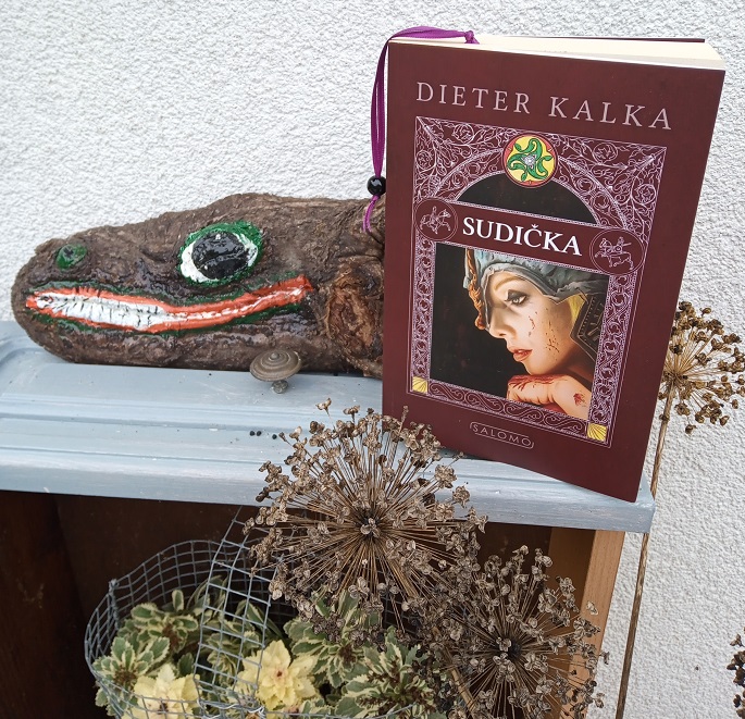Das Buch steht auf einer Schublade, die mit Pflanzen und einem bemalten Holzstück dekoriert ist.