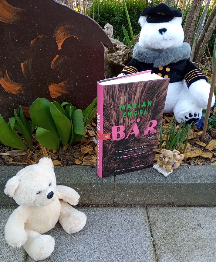 Das Buch steht in einem Blumenbeet, umrahmt von zwei Plüschbären und einem Keramikbären, der bäuchlings auf einem aufgeschlagenen Buch liegt.