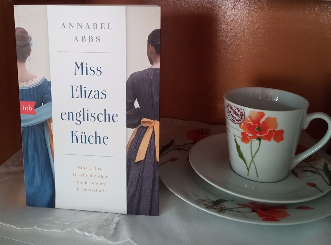 Ein Buch steht in einem Küchenschrank, der mit einer Decke ausgelegt ist. Daneben steht ein Gedeck: Teller, Untertasse, Tasse mit Blumenmuster