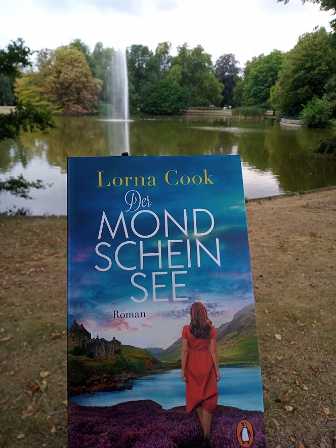 Ich halte das Buch hoch, im Hintergrund  sieht man eine Fontäne in einem See.