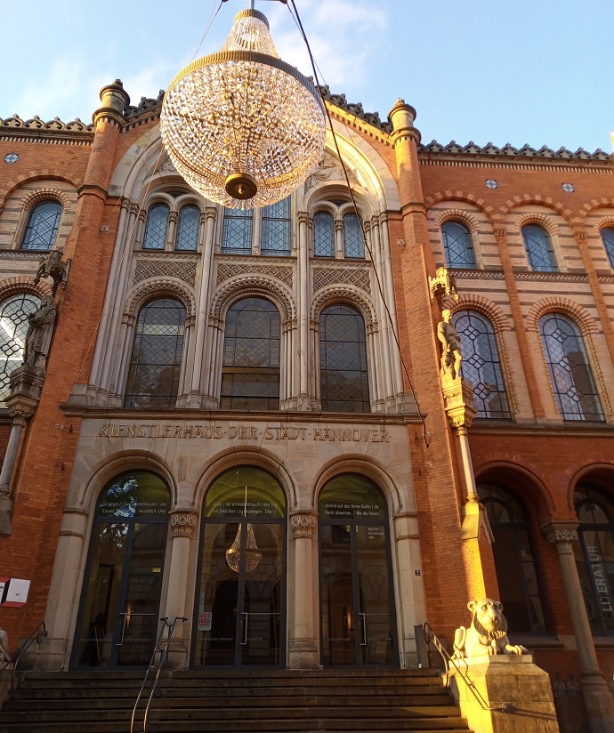 Das gleiche Gebäude wie vorher, nur bei Tag und nicht die komplette Front im Bild zu sehen.
Vor dem Gebäude hängt ein großer Kronleuchter über der Straße.
Über den Eingangsportalen steht: Künstlerhaus der Stadt Hannover