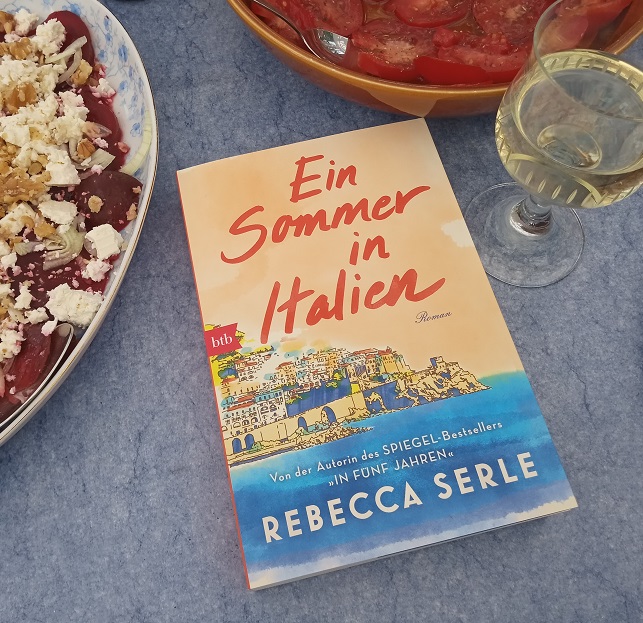 Das Buch liegt auf einem Tisch neben einer Schale Tomaten, einer Schale rote Beete mit Schafskäse und einem Glas Weißwein