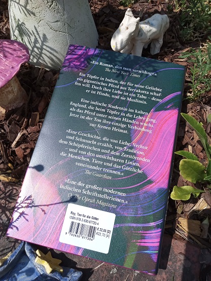 Das Buch liegt mit der Rückseite nach oben auf einer Schicht Rindenmulch, darüber steht ein kleines getöpfertes Pferd.