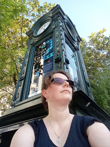 Ein Selfie. Ich sitze vor der Kröpcke-Uhr, im Hintergrund sind Bäume zu sehen. Ich trage eine Sonnenbrille, denn es ist ein sehr sonniger Septembertag. Viel zu warm für diese Jahreszeit.