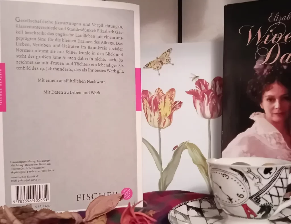 Das Buch steht (mit dem Klappentext nach vorne) neben einer Blumen-Postkarte, davor liegt eine getrocknete Rose, daneben steht ein kleiner Alice im Wunderland Eierbecher. Es ist ein Teil der DVD am Bildrand zu sehen.